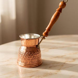 May include: A copper coffee pot with a silver-toned interior and a long, turned wooden handle. The exterior of the pot features an embossed scale pattern and geometric detailing around the base and neck, with a small metal loop at the handle end.