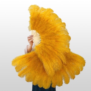 May include: A large, vibrant yellow ostrich feather fan featuring long, soft plumes attached to a light-coloured wooden frame. The fan is fully extended, showcasing a wide, semi-circular shape with dense, fluffy feathers that create a voluminous texture.