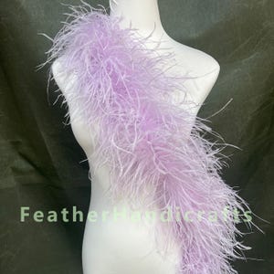 Puede incluir: Una boa de plumas de color lavanda claro drapeada en diagonal sobre un torso de maniquí blanco. Las plumas largas, suaves y vaporosas crean una textura voluminosa. El texto FeatherHandicrafts está impreso en verde claro en la imagen.