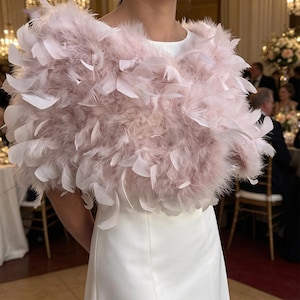 May include: A pale pink feather shrug worn over a plain white sleeveless dress. The garment features a dense layer of soft, fluffy down feathers interspersed with longer, structured feathers, creating a voluminous and textured appearance.