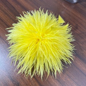 May include: A bright yellow feather accessory shaped like a round, dense pom-pom. The object consists of numerous thin, radiating feathers that create a full, spherical appearance with a vibrant, uniform color throughout the entire structure.