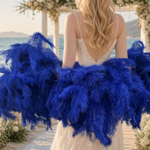 May include: A vibrant royal blue ostrich feather shrug worn over a white sleeveless dress with a V-neckline. The voluminous, fluffy feathers create a wide, textured wrap that covers the shoulders and upper arms, providing a bold contrast to the dress.