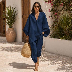 May include: A navy blue linen two-piece outfit. The top has a V-neck and wide sleeves. The pants are a relaxed fit. A woven tote bag is carried. The outfit is worn with sandals and sunglasses.