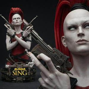 May include: A collectible bust of the character Aurra Sing featuring a pale-skinned figure with a tall red ponytail, wearing a red top and black wristbands. The figure holds two metallic blasters. The base displays the text AURRA SING.