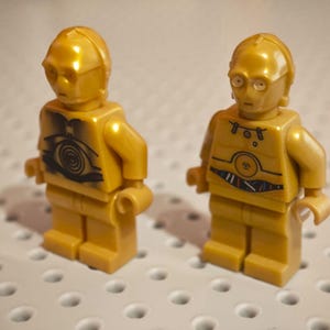 Könnte beinhalten: Zwei goldfarbene Lego-Figuren der C-3PO-Figur aus Star Wars. Jede Figur hat ein detailliertes Design auf dem Torso und eine markante Kopfform. Die Figuren stehen auf einer weißen Oberfläche.
