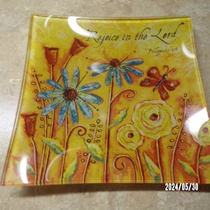 May include: Square glass dish with a yellow background and a floral design. The flowers are blue, yellow, and orange. There is a butterfly in the upper right corner. The text "Rejoice in the Lord" is written in a cursive font at the top of the dish. The text "Philippians 4:4" is written in a smaller font below the main text.