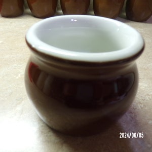 May include: A small, brown ceramic pot with a white rim. The pot is round and has a smooth, glossy finish.