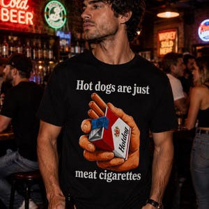 Könnte beinhalten: Ein schwarzes T-Shirt mit einem Grafikdruck einer Hand, die eine rot-weiße Zigarettenschachtel mit der Aufschrift Hotdog hält. Der Text über der Grafik lautet Hot dogs are just und der Text unter der Grafik lautet meat cigarettes.