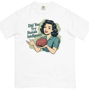 May include: A white short-sleeve t-shirt featuring a graphic illustration of a person with dark hair wearing a teal shirt, holding a pink brain. The text "Did You Try Human Intelligence?" is printed in dark teal next to the illustration.