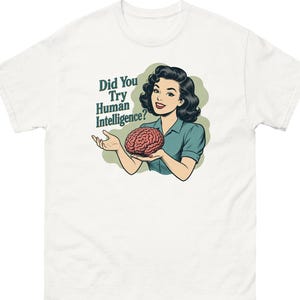 May include: A white t-shirt featuring a graphic illustration of a person with dark hair wearing a teal shirt, holding a pink brain in their hands. The text "Did You Try Human Intelligence?" is printed in dark green letters next to the illustration.