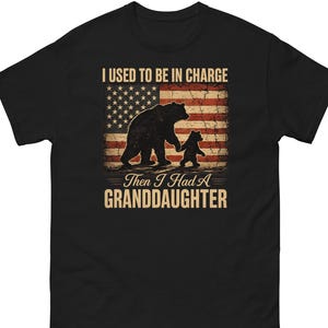 May include: Black t-shirt featuring a distressed American flag design with a bear and cub silhouette. The text reads "I USED TO BE IN CHARGE, Then I Had A GRANDDAUGHTER".