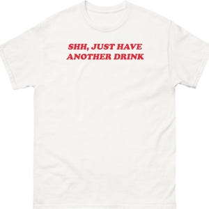 May include: A white short-sleeved t-shirt featuring the red text SHH, JUST HAVE ANOTHER DRINK printed in a bold, sans-serif font across the chest. The garment has a crew neckline and a standard fit, presenting a clean and simple design.