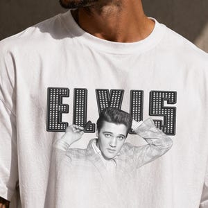 May include: A white short-sleeved t-shirt featuring a black and white graphic of Elvis Presley with the text ELVIS in a bold, dotted font above the image. A small tag attached to the shirt displays the text The Gifted Tee Lab Purely Crafted Tees.