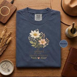 May include: A navy blue t-shirt with a floral design featuring white and peach flowers, green leaves, and the text "Be your own kind of beautiful." The shirt is surrounded by accessories on a wooden surface.