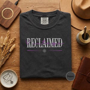 May include: Pepper-colored t-shirt with the word "RECLAIMED" in white and purple script. The shirt has a crew neck and a Comfort Colors tag. The shirt is displayed on a wooden surface with accessories.