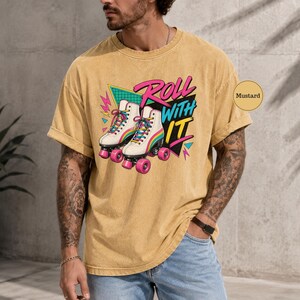 A man wearing a yellow t-shirt with a graphic of roller skates and the text "Roll with it" is standing in front of a concrete wall.