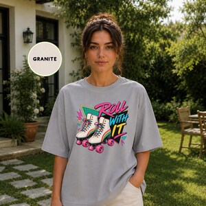 A woman wearing a gray t-shirt with a graphic design of rollerblades and the text "Roll with it" stands in front of a house with a sign that says "Gifted Tee Lab".