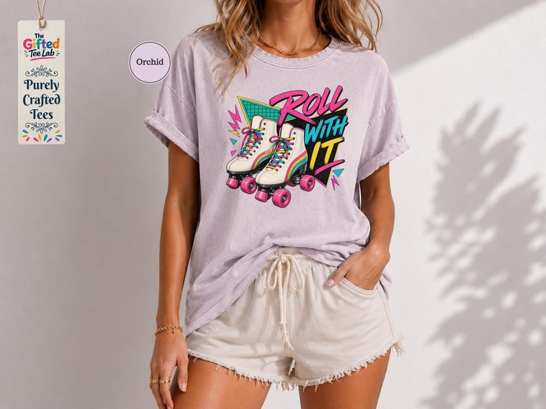 A woman wearing a pink t-shirt with a graphic design of rollerblades and the text "Roll with it" is standing in front of a white wall.