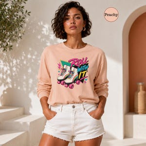 A woman wearing a pink long-sleeved shirt with a graphic of roller skates and the text "Roll with it" stands in front of a pink wall with a tree and a vase.
