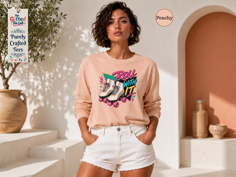 A woman wearing a pink long-sleeved shirt with a graphic of roller skates and the text "Roll with it" stands in front of a pink wall with a tree and a vase.