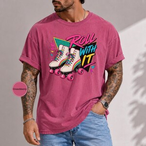 A man wearing a pink t-shirt with a graphic of roller skates and the text "Roll with it" is standing in front of a gray wall.