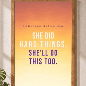 Op de afbeelding: Een rechthoekige wandkunstprint in een houten lijst met een zonsondergang-geïnspireerde gradiëntachtergrond die verloopt van oranje naar geel. De tekst luidt: - for the woman who keeps going - SHE DID HARD THINGS. SHE'LL DO THIS TOO.