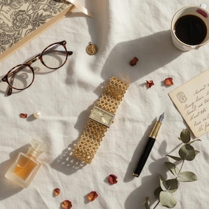 Può includere: Un orologio da polso dorato con quadrante rettangolare e cinturino in maglia testurizzata, accompagnato da occhiali tartarugati, una penna stilografica, un piccolo flacone di profumo, un ciondolo d'oro, un libro, una tazza di caffè e petali.