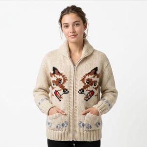 May include: Cream-colored zip-up cardigan sweater with a shawl collar and two front pockets. The sweater features a repeating pattern of deer faces in brown, black, and red. Blue floral accents are on the sleeves and pockets.