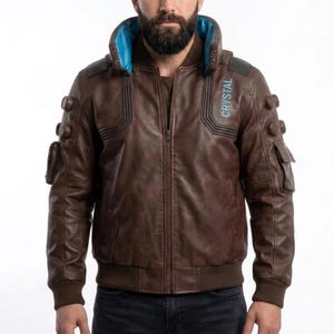 Puede incluir: Chaqueta de cuero marrón con cuello azul y la palabra "CRYSTAL" impresa en el hombro derecho. La chaqueta tiene múltiples bolsillos y puños de canalé. Es una chaqueta estilo bomber.