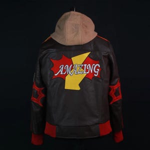 Side and rear angle view of a Marvel Rivals Inspired Spider-Man Bomber Leather Jacket with Hoodie, showcasing a large embroidered red comic book style burst patch on the back with the word "AMAZING" written in white across a yellow lightning bolt. The sleeve displays red web embroidery and yellow and red shoulder stripe details.