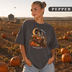 May include: A charcoal gray t-shirt featuring a graphic of a skeleton wearing a witch hat, sitting in a wooden wheelbarrow filled with pumpkins. The design includes a crescent moon, bats, and the text PUMPKIN PICKING CREW in white lettering.