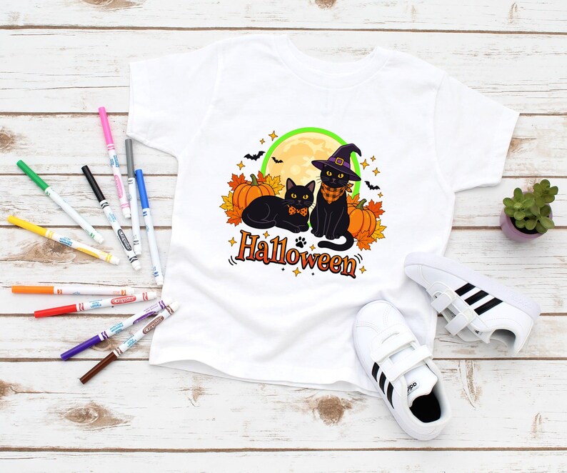 A graphic design featuring two black cats, one wearing a purple witch hat and orange bandana, sitting among pumpkins and autumn leaves under a full moon with the word Halloween below.