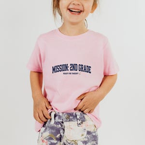 May include: A light pink short-sleeved t-shirt featuring the dark blue text MISSION: 2ND GRADE and READY FOR TAKEOFF with a small rocket ship icon. The shirt is paired with blue and white floral patterned denim shorts with a visible metal button.