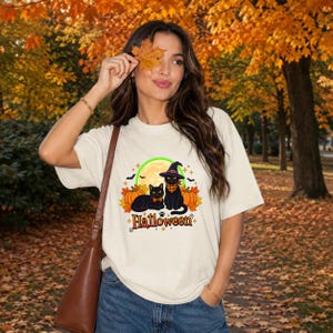 A graphic design featuring two black cats, one wearing a purple witch hat and orange bandana, sitting among pumpkins and autumn leaves under a full moon with the word Halloween below. — shown on a ivory product