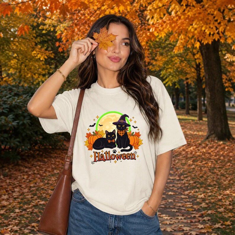 A graphic design featuring two black cats, one wearing a purple witch hat and orange bandana, sitting among pumpkins and autumn leaves under a full moon with the word Halloween below. — shown on a ivory product