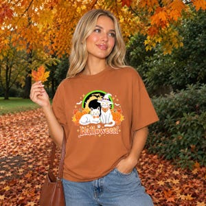 A graphic design featuring two black cats, one wearing a purple witch hat and orange bandana, sitting among pumpkins and autumn leaves under a full moon with the word Halloween below.
