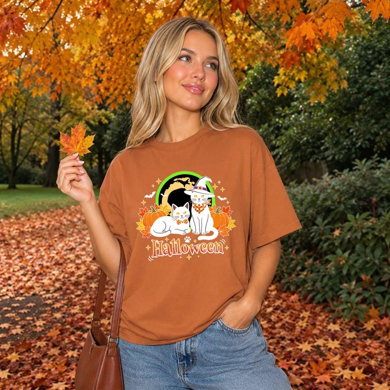 A graphic design featuring two black cats, one wearing a purple witch hat and orange bandana, sitting among pumpkins and autumn leaves under a full moon with the word Halloween below.