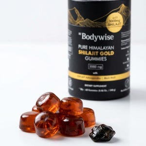 Pode incluir: Um recipiente cil&iacute;ndrico preto com gr&aacute;ficos de montanhas douradas e texto para "Bodywise Pure Himalayan Shilajit Gold Gummies" est&aacute; ao lado de uma pilha de gomas de cor &acirc;mbar e um peda&ccedil;o escuro de forma irregular.