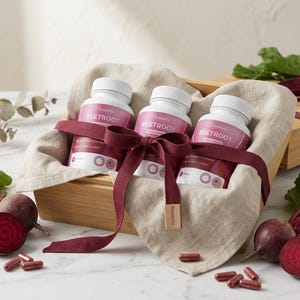 May include: Three white bottles labeled "Beetroot" are nestled in a wooden box lined with beige fabric, tied with a burgundy ribbon. Fresh beets and capsules are arranged around the box, suggesting a health and wellness theme.