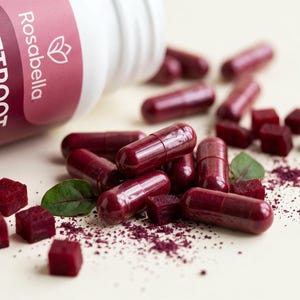 May include: A white bottle of Rosabella Beetroot capsules is tipped over, spilling out several capsules, beetroot cubes, and beetroot powder. The capsules are a deep red color, and the cubes are a vibrant red. Green leaves are scattered around.