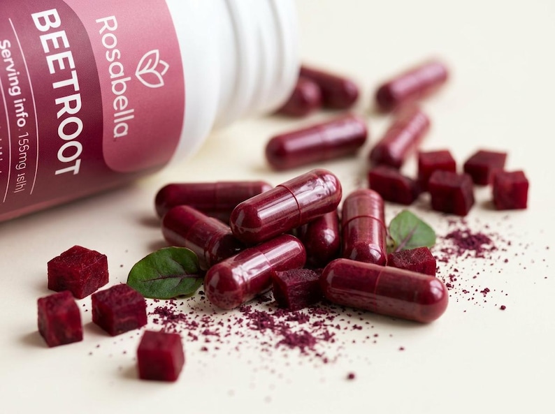 May include: A white bottle of Rosabella Beetroot capsules is tipped over, spilling out several capsules, beetroot cubes, and beetroot powder. The capsules are a deep red color, and the cubes are a vibrant red. Green leaves are scattered around.