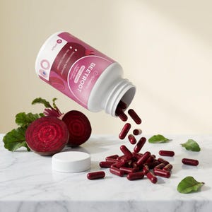Puede incluir: Una botella blanca de c&aacute;psulas de remolacha con una etiqueta rosa, derram&aacute;ndose sobre una superficie de m&aacute;rmol. La etiqueta dice "Beetroot" y "Rosebella". Tambi&eacute;n hay remolacha en rodajas y hojas verdes, junto con la tapa blanca de la botella.