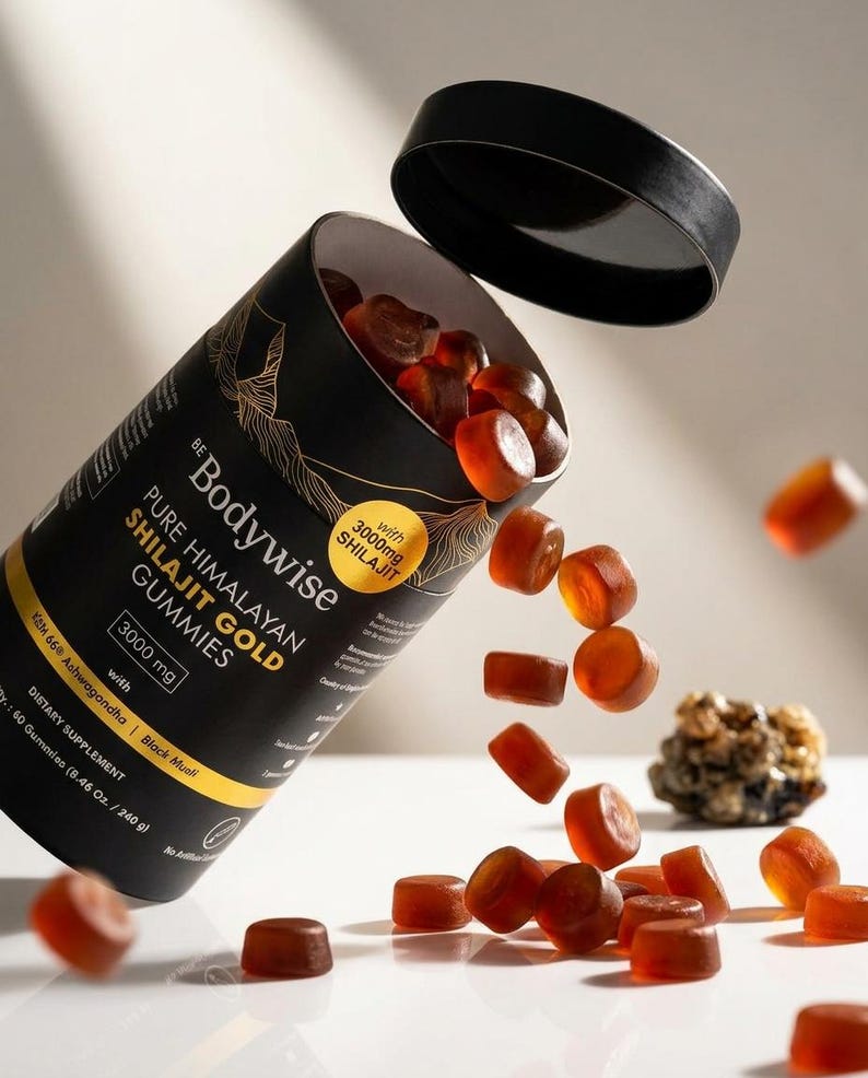 Pode incluir: Um recipiente cil&iacute;ndrico preto de Bodywise Pure Himalayan Shilajit Gold Gummies com a tampa aberta, e gomas a sair. O recipiente tem letras douradas e um desenho de montanha dourada. As gomas s&atilde;o de cor &acirc;mbar transl&uacute;cida.