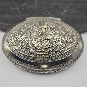 May include: An oval-shaped silver-toned metal box featuring an intricate embossed design of a bird on the lid. The surface is decorated with detailed scrollwork and textured patterns, with a small hint of gold-toned metal visible on the bird.