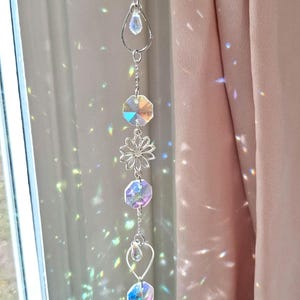 Puede incluir: Un atrapasoles vertical colgante que presenta una cadena de metal plateado con cristales de vidrio facetados transparentes alternados y un dije de flor plateado central. Los cristales reflejan la luz, creando un efecto de prisma de arcoíris.