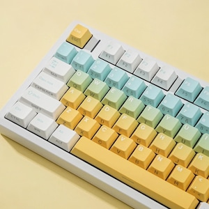 May include: A mechanical keyboard featuring a white frame and a gradient keycap set. The keys transition from light blue at the top left to soft green in the center and warm yellow at the bottom, with white function and modifier keys.