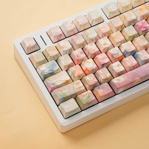 May include: A mechanical keyboard featuring a white frame and custom keycaps with a soft, multi-colored floral watercolor pattern. The keys display standard alphanumeric characters, function keys, and command labels in a clear, white sans-serif font.