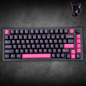 Könnte beinhalten: Eine mechanische Tastatur mit dunkelgrauem Rahmen und Tasten, die mit rosa Beschriftung und Akzenten versehen sind. Das Layout umfasst einen Drehknopf oben rechts, eine rosa Leertaste sowie spezielle rosa Tasten mit Symbolen für Smiley, Ahornblatt und Kartensymbole.