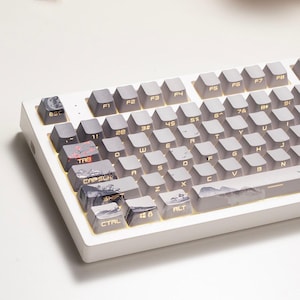 May include: A white mechanical keyboard featuring grey keycaps with gold lettering. Several keycaps on the left side and the spacebar display a printed landscape illustration with cherry blossoms and mountains. The keys include standard labels.