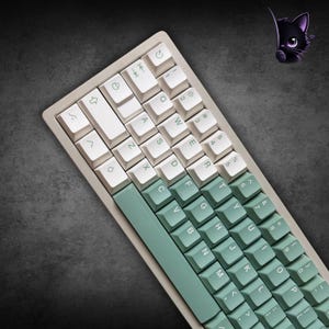 May include: A mechanical keyboard featuring a two-tone colour scheme with white keys on the left and sage green keys on the right. The keys display standard alphanumeric characters, symbols, and function icons in a dark green font. The keyboard frame is a light beige colour.